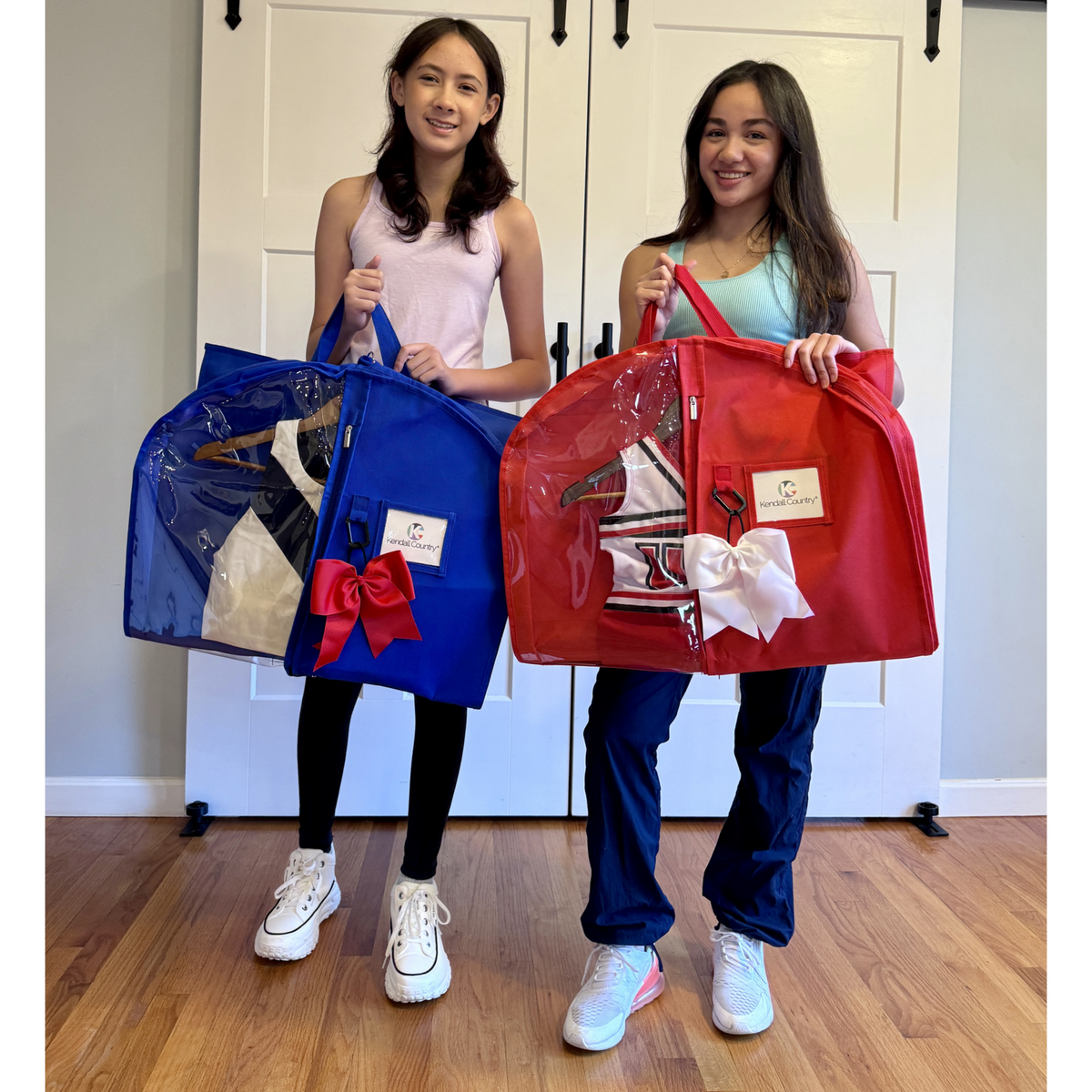 Two girls standing indoors holding large zippered garment bags. The bag on the left is royal blue with a red bow, and the bag on the right is bright red with a white bow.