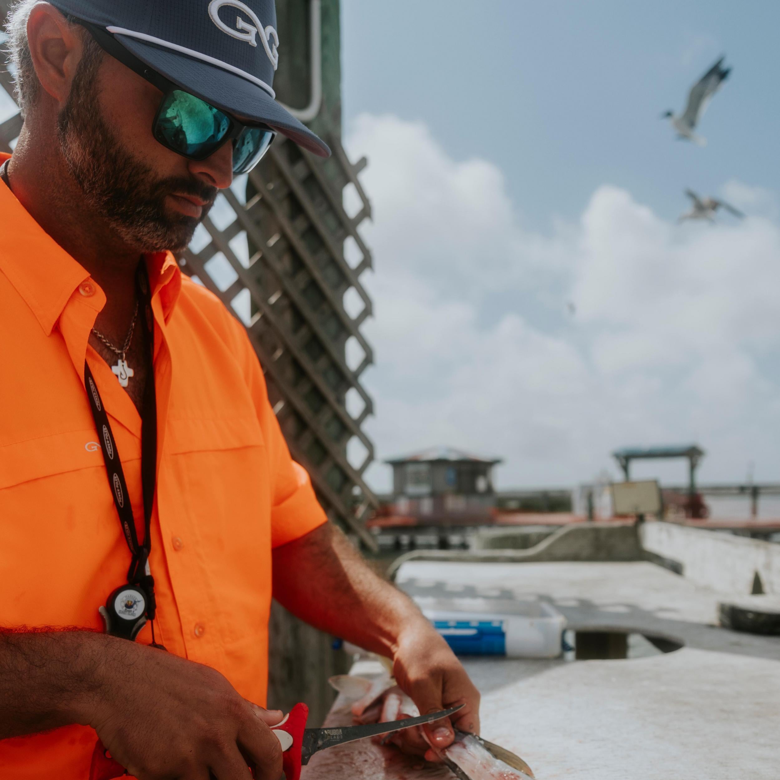 Man in orange shirt and cap with sunglasses holding a knife on a rooftop with seagulls and a building in the background.