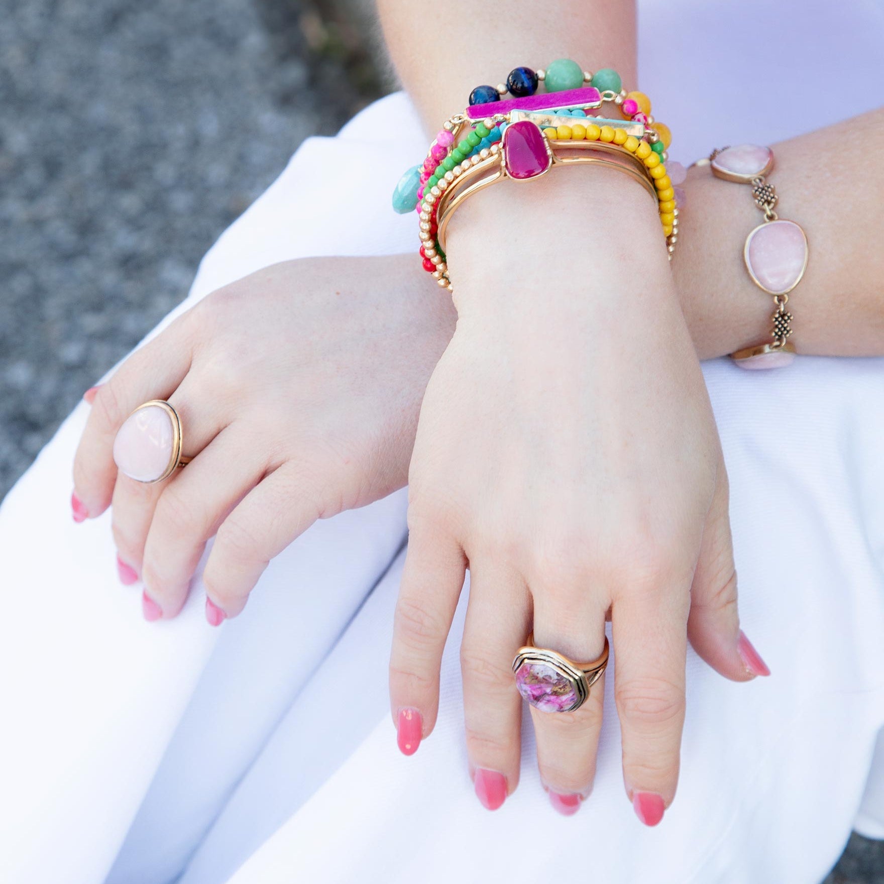A close-up of a person's hands wearing multiple pieces of jewelry, including colorful beaded bracelets, a pink stone ring, and a pink stone bracelet, resting on white fabric.