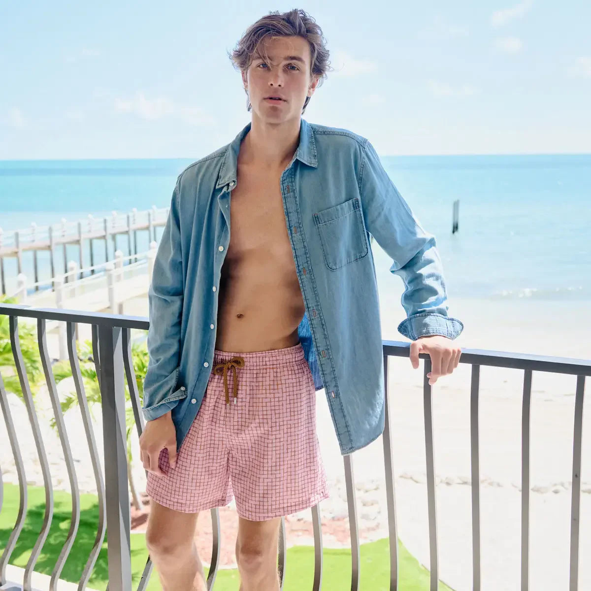 Man wearing a blue shirt over a tank top and pink shorts on a balcony with a beach view.