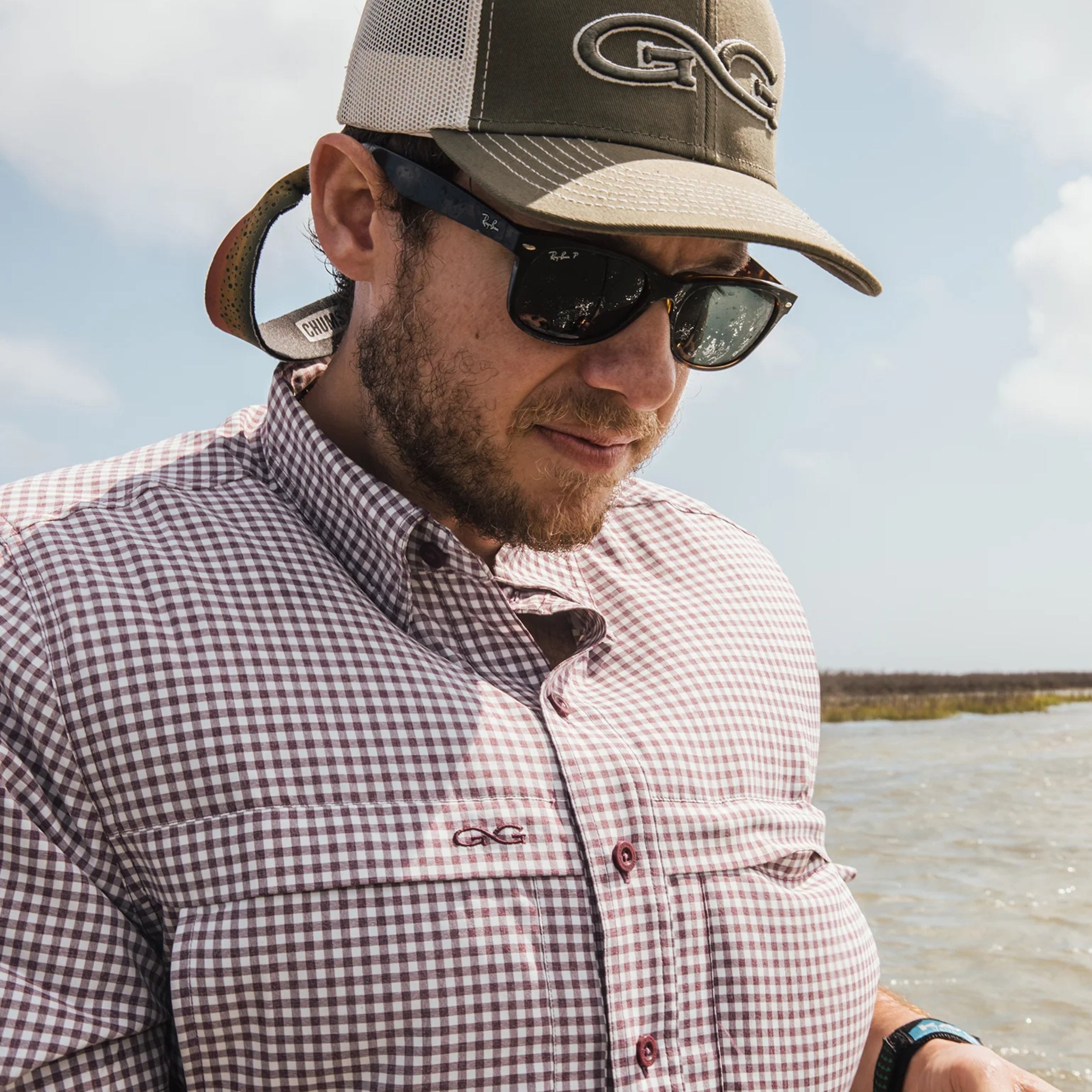 Man wearing a checkered shirt and cap with a logo, standing by a body of water.