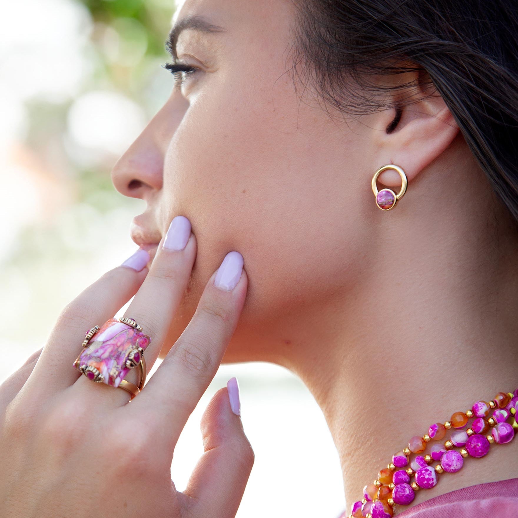 Woman wearing pink and gold jewelry including earrings, ring, and necklace with blurred greenery in the background