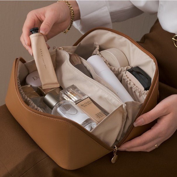 Person organizing cosmetic products in a brown travel bag.