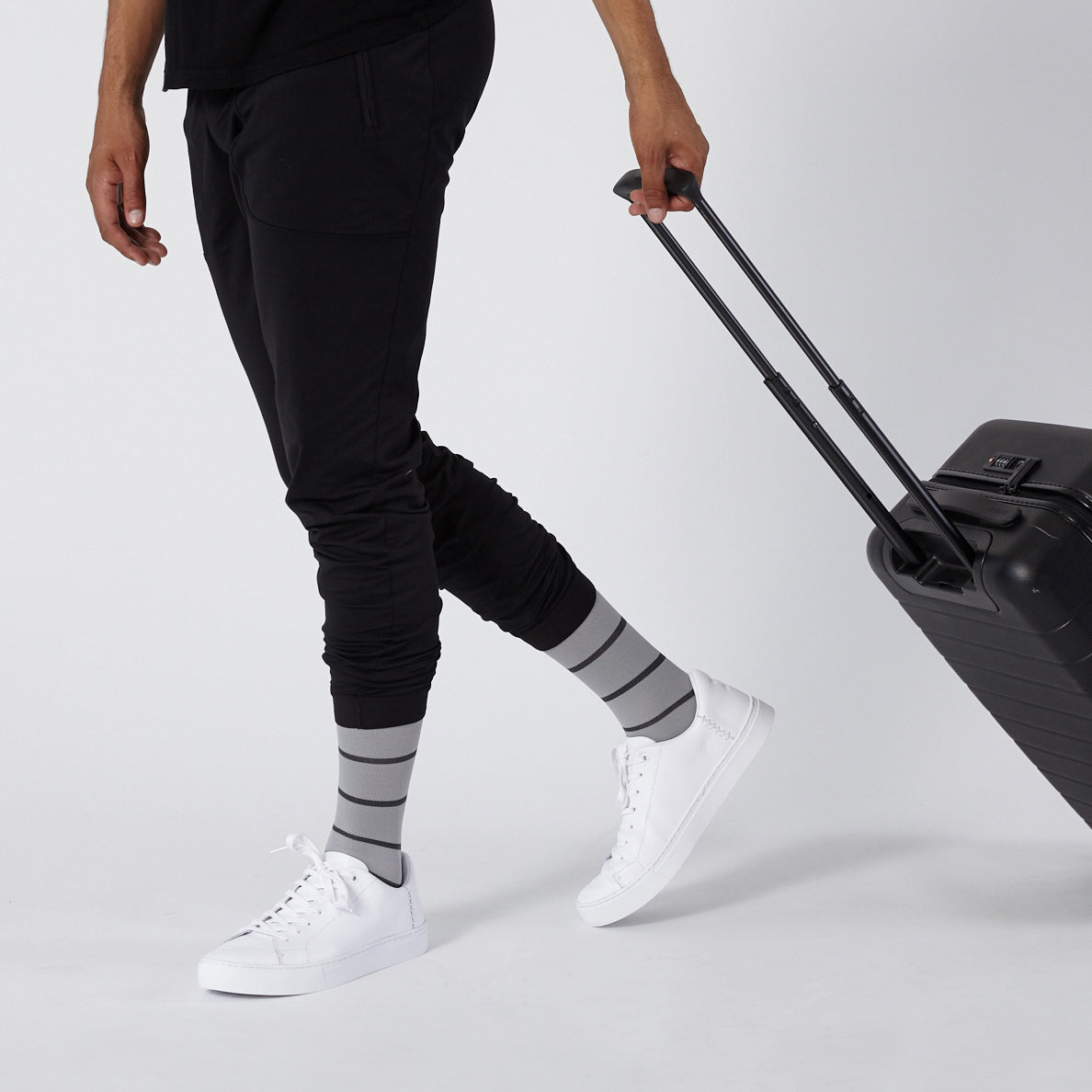Person wearing black pants, gray striped socks, and white sneakers with a suitcase on a light background
