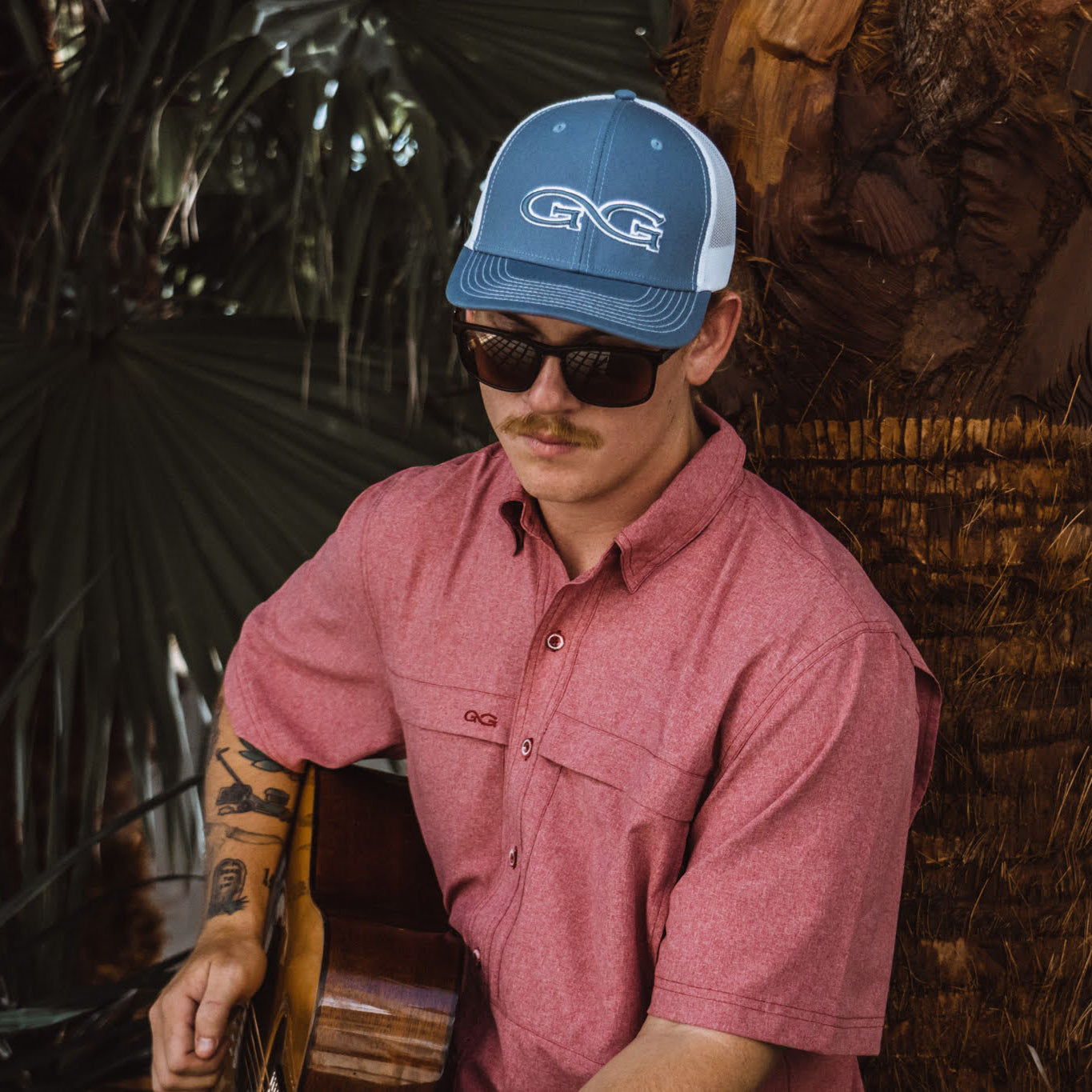 Man wearing a blue cap and sunglasses, sitting against a natural background