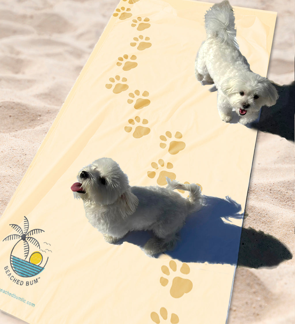 Two small white dogs on a beach mat with paw prints, brand logo visible.