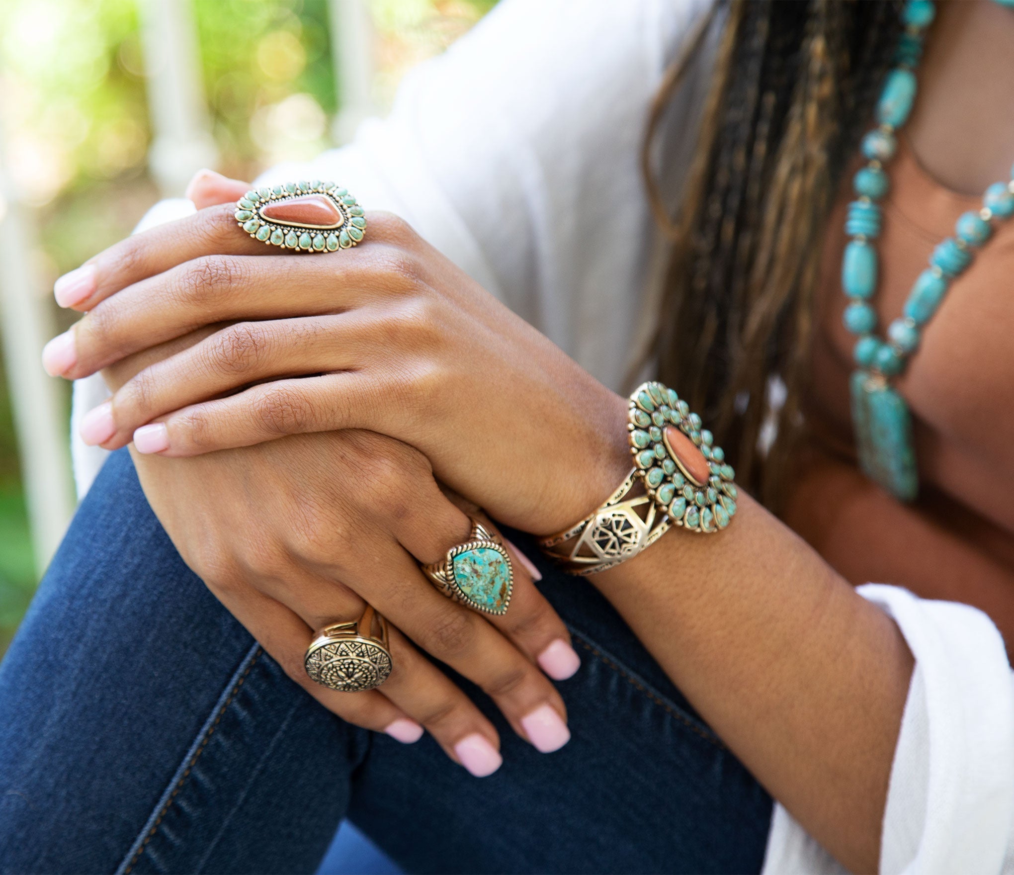 Close-up of a person's hands wearing multiple turquoise rings and a bracelet, with a blurred natural background.