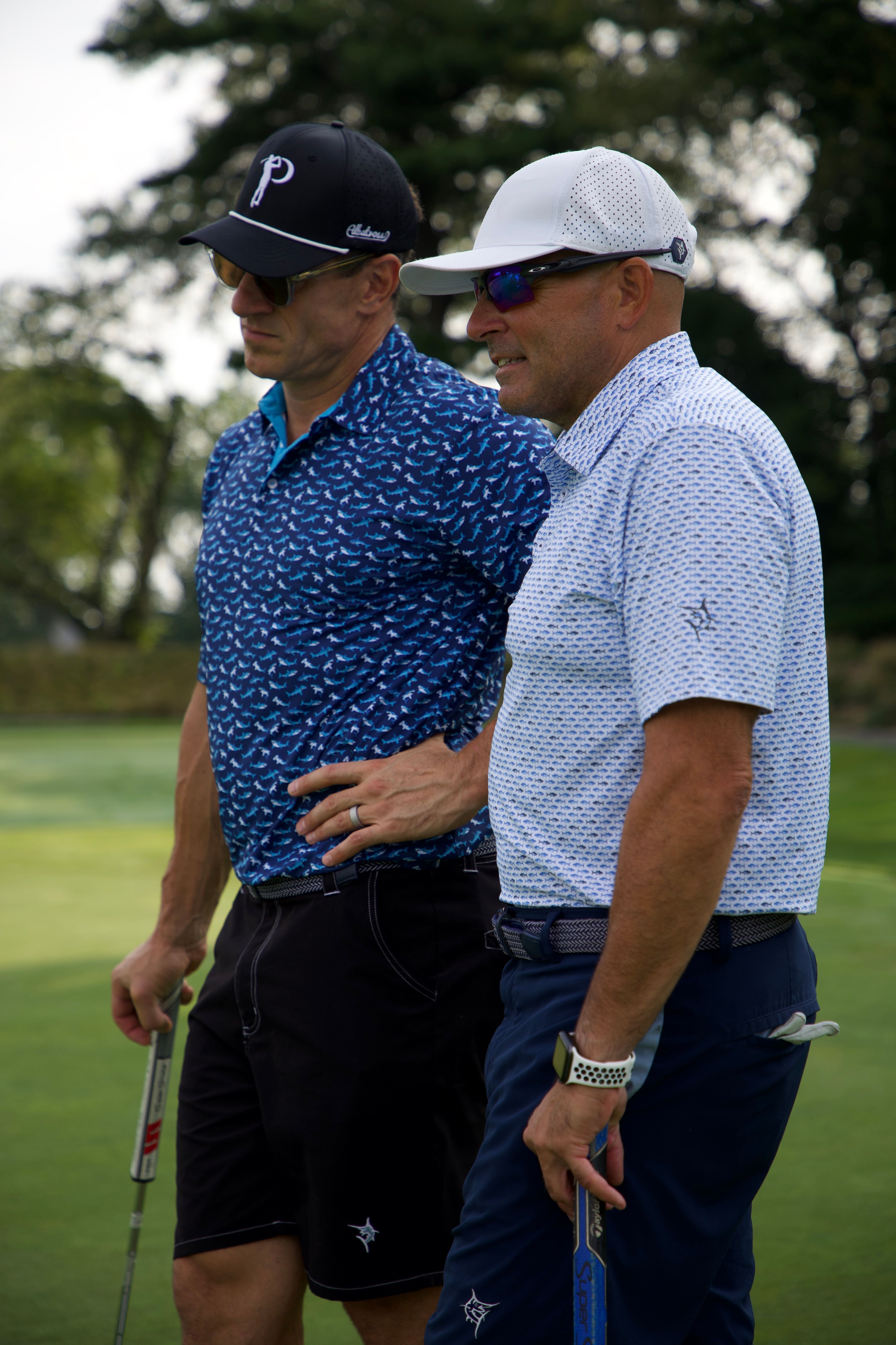 Two men on a golf course, one wearing a blue patterned shirt and black cap, the other in a light blue shirt and white cap.