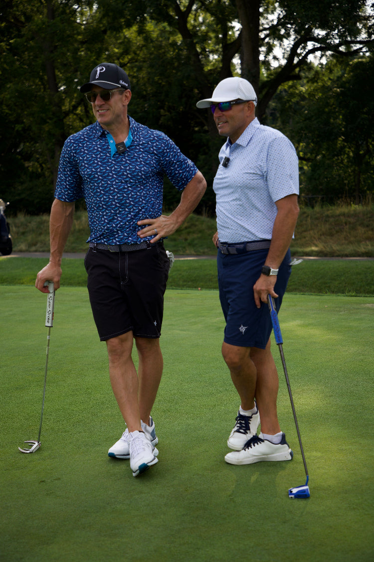 Two men on a golf course with golf clubs, wearing sunglasses and casual attire.