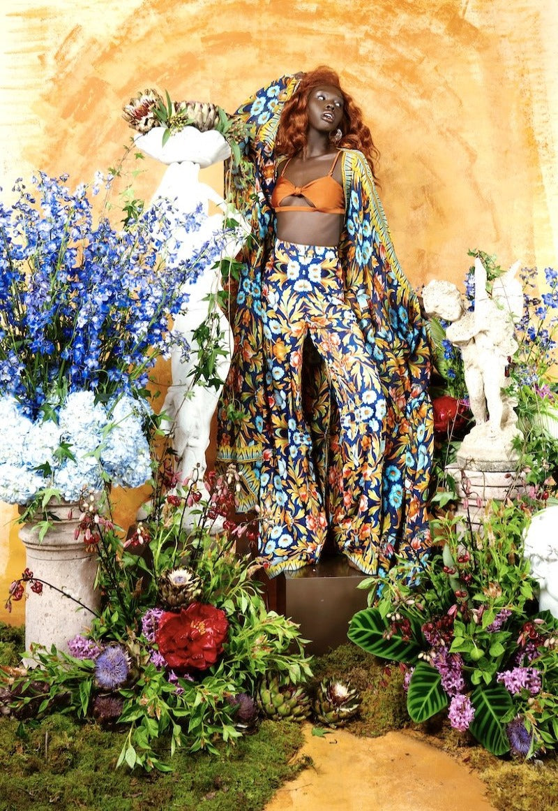 Person in a colorful floral outfit surrounded by flowers and statues against a yellow wall.