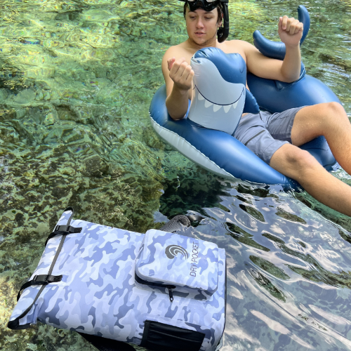 Person on a shark-shaped inflatable tube in clear water with a camouflage Dri-Pac bag nearby.