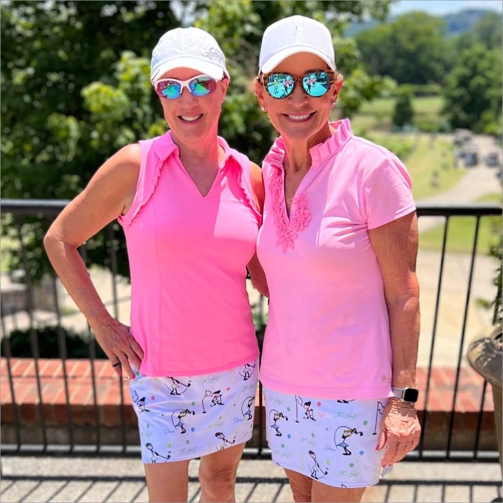 Two women in pink shirts and white hats standing outdoors with a scenic background.