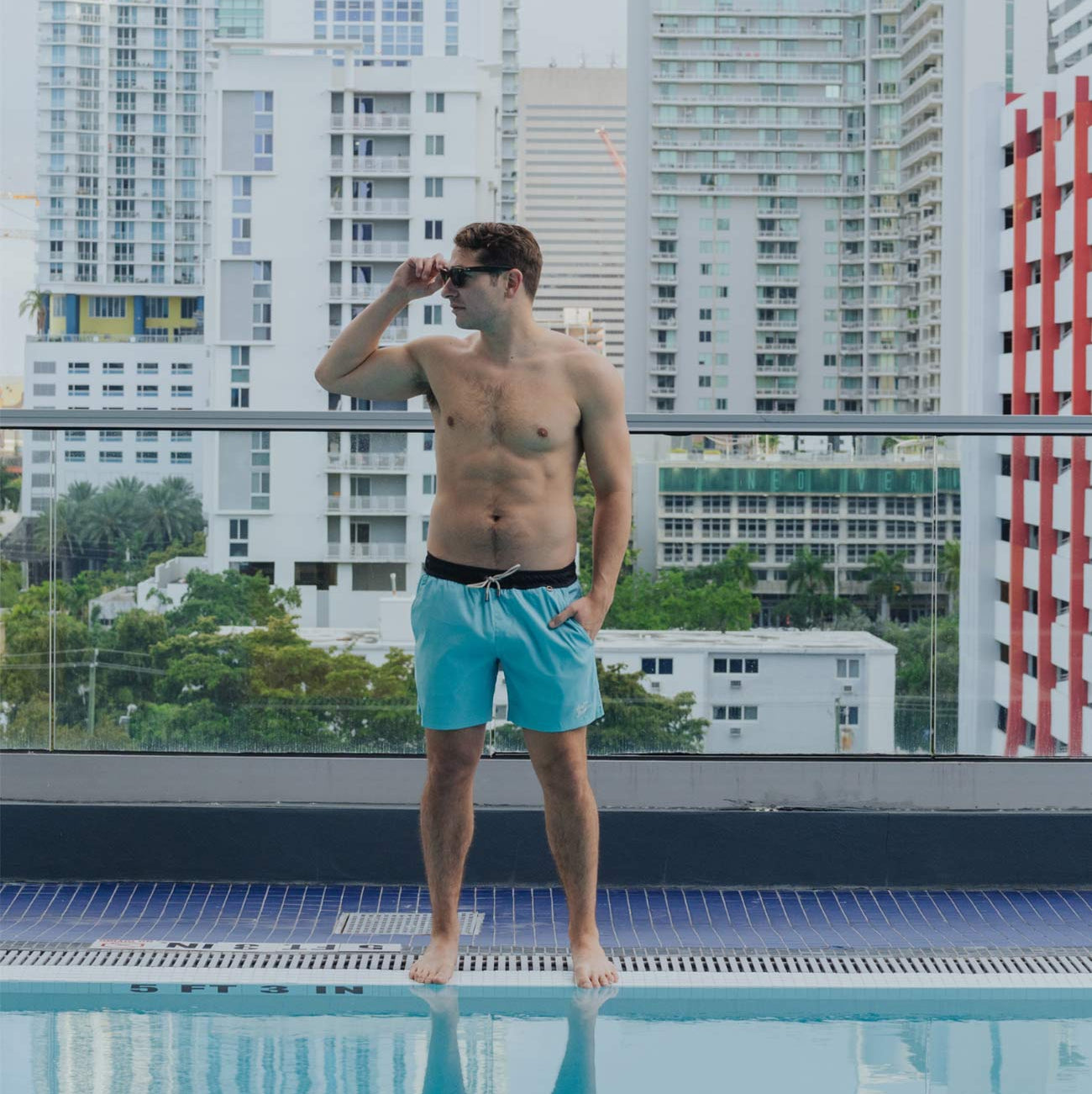 Man in turquoise shorts standing by a pool with city skyline in the background