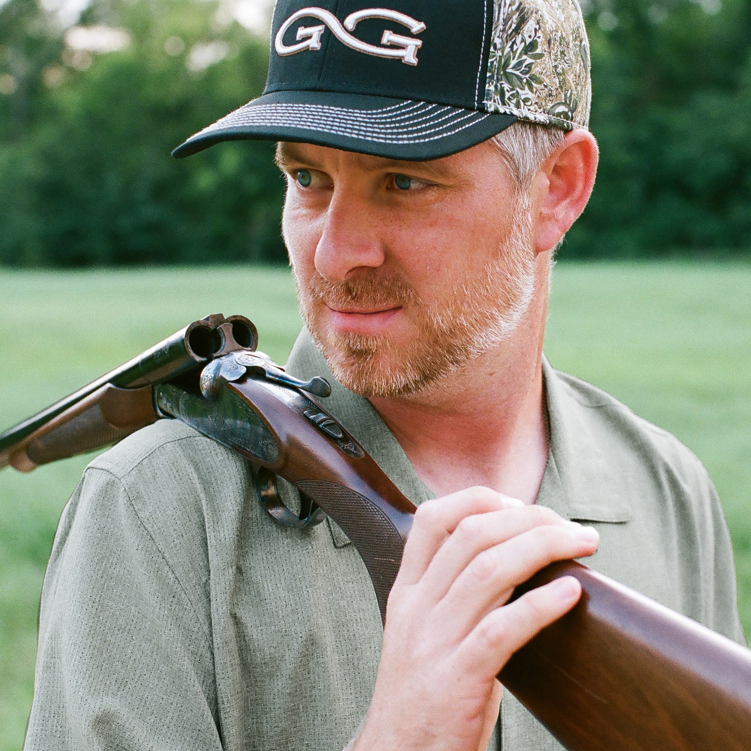 A man outdoors wearing a black and camouflage baseball cap with a white GG logo and holding a shotgun over his shoulder.