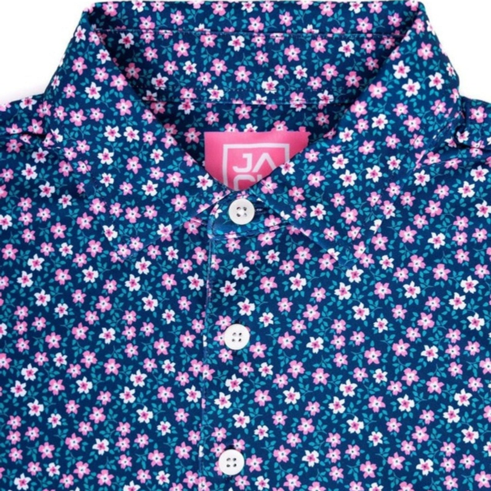 A close-up of a short-sleeve button-up shirt with a dark teal blue background covered in a dense pattern of small pink and white flowers. The shirt features a classic collar, white buttons, and a pink interior neck label with a white logo.