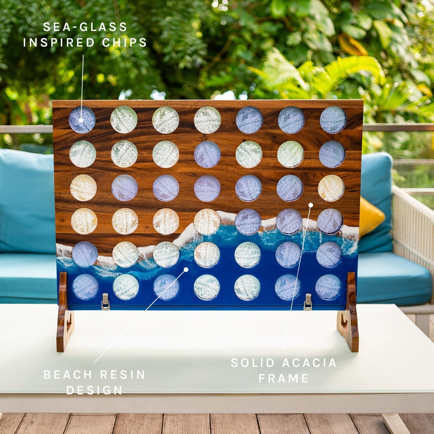 A decorative rectangular wooden board with a grid of circular cutouts showing beach-inspired resin designs in blue, white, and gray, displayed on a table outdoors. Text labels point to sea-glass inspired chips, beach resin design, solid acacia frame, and the overall design.