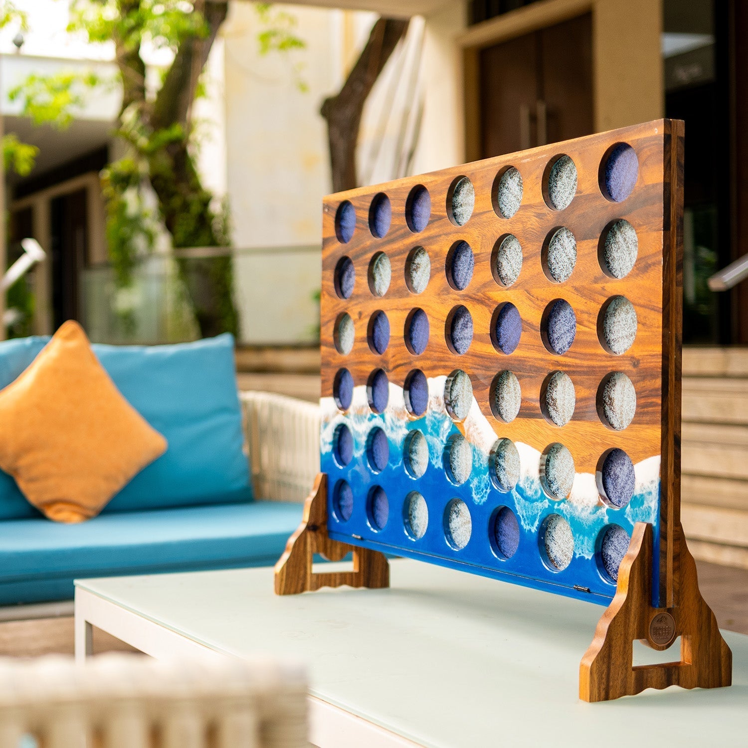 A wooden Connect 4 game board with a blue ocean wave design near the bottom, displayed on a white table in a modern patio setting with a blue sofa in the background.