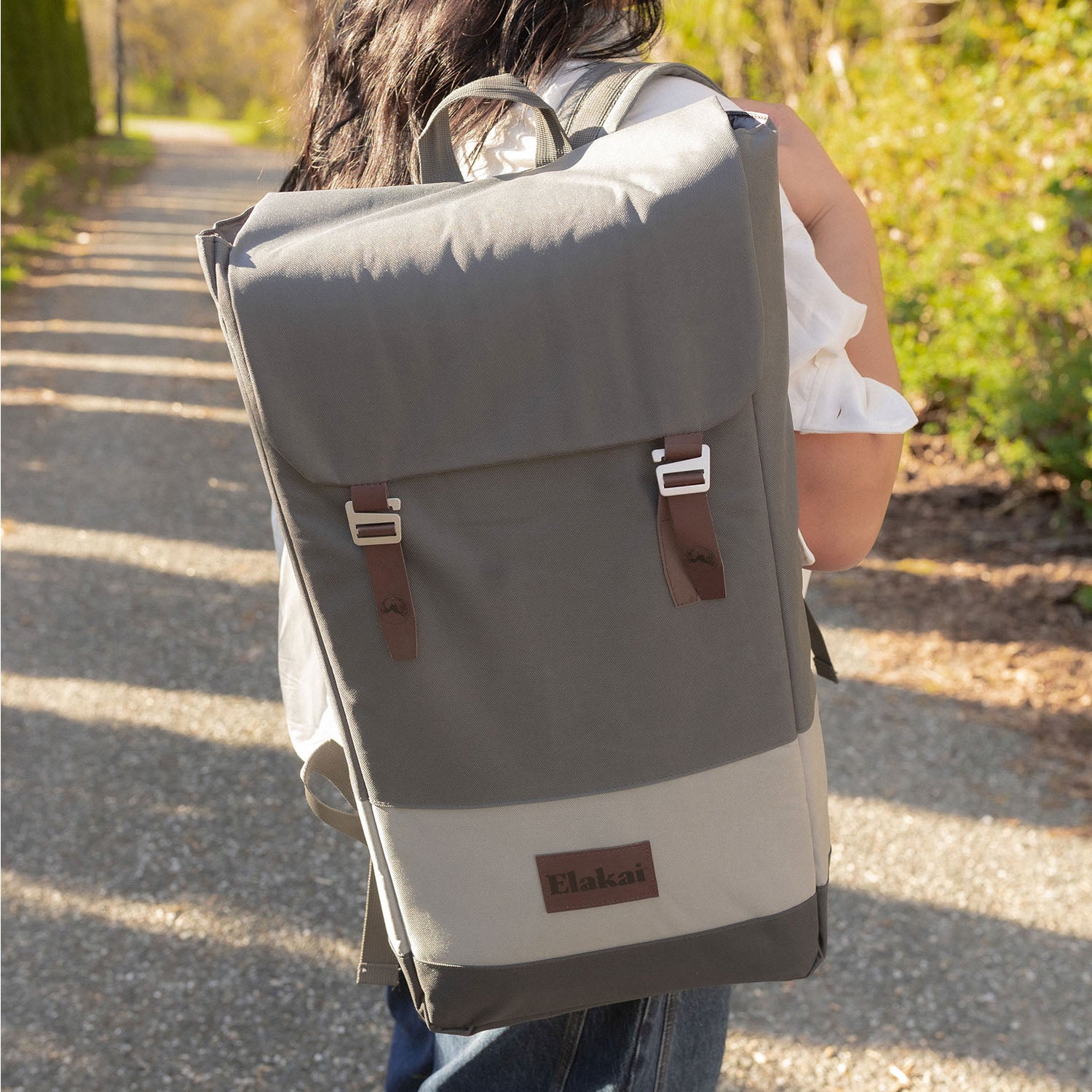 A person viewed from behind wearing a gray and off-white backpack with brown leather-look straps and a small 'Elokai' brand patch, walking on a sunlit path outdoors.
