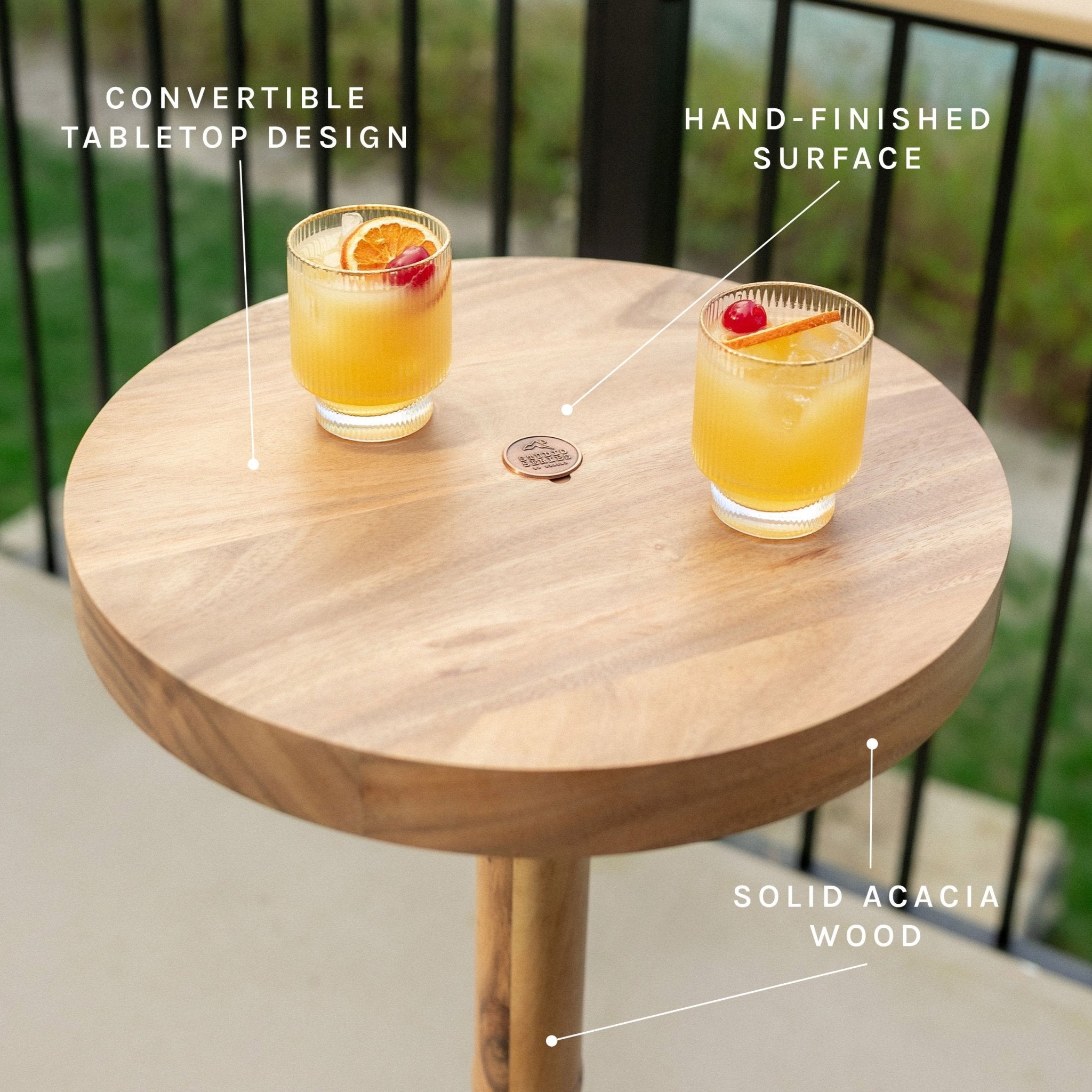 Round solid acacia wood convertible tabletop with two drinks on top, shown outdoors with text callouts reading 'Convertible tabletop design,' 'Hand-finished surface,' and 'Solid acacia wood.'.