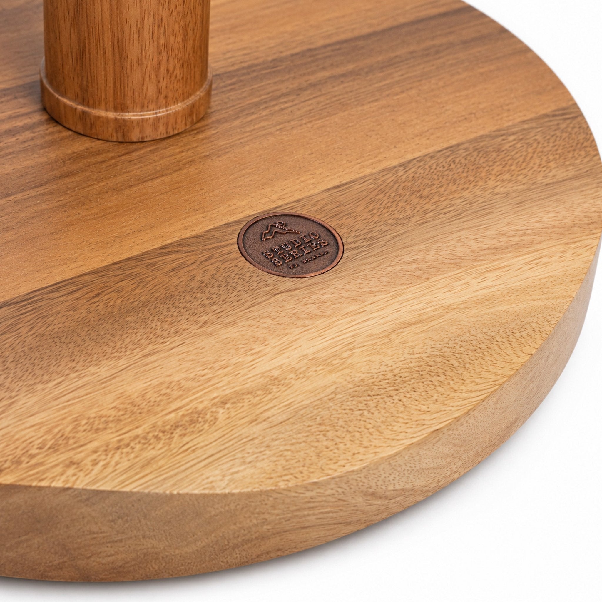 Close-up of a round wooden table base with a visible metal brand plaque reading 'BETTER BENCH' and a cylindrical wooden leg attached at the top left, shown on a white background.