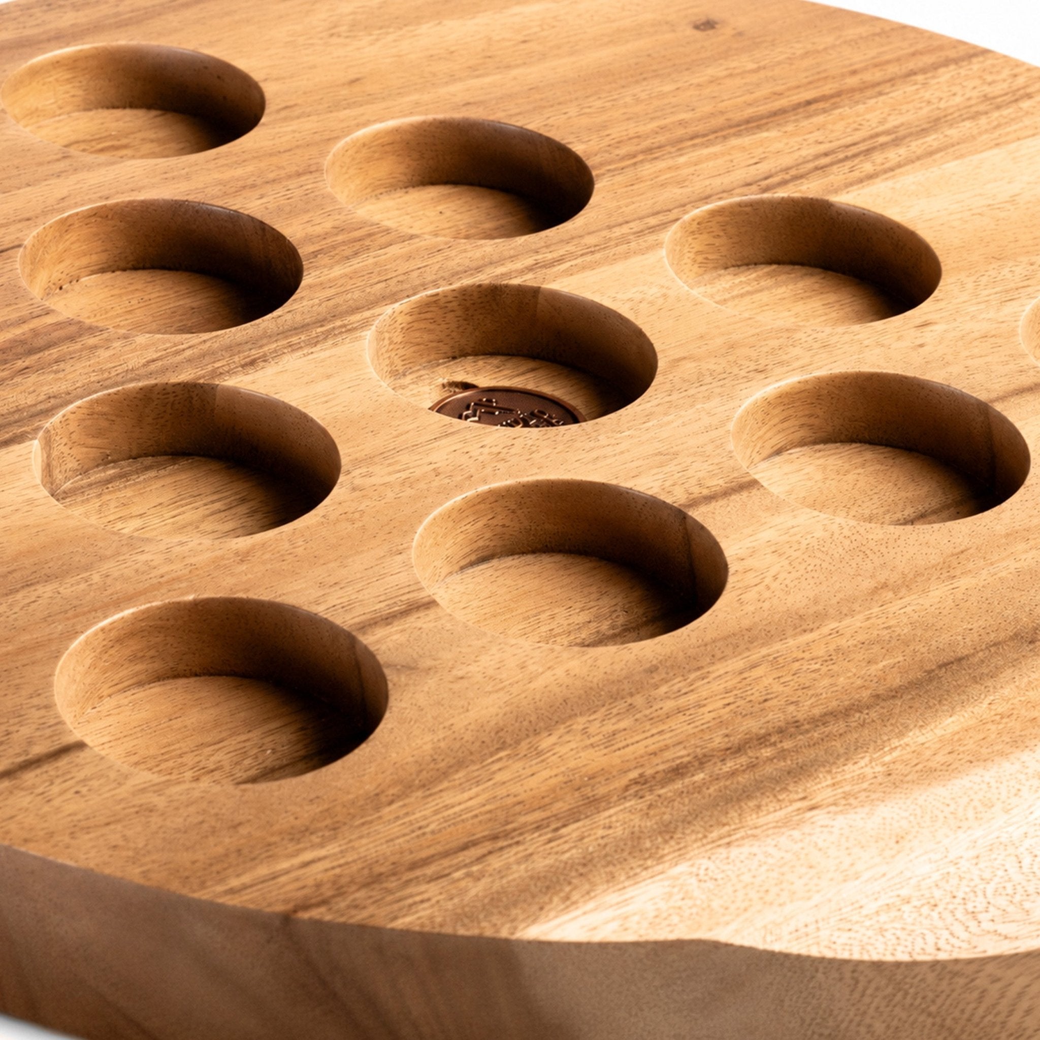 Close-up of a round wooden serving board with eight circular recessed wells, showing natural wood grain and a small dark food piece in one well.