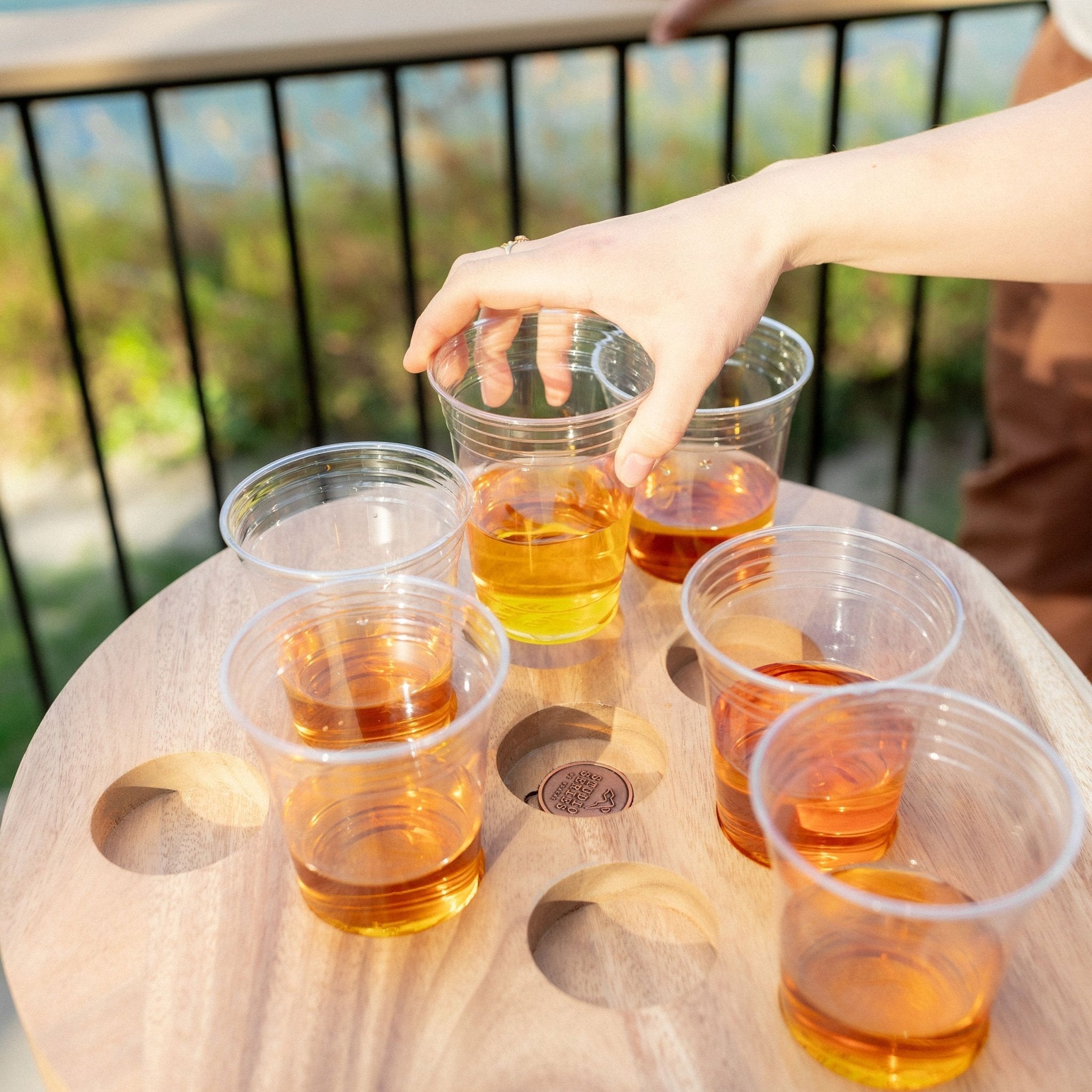 A hand holding a clear plastic cup filled with amber-colored liquid, set on a round wooden tray with several similar cups in an outdoor setting.