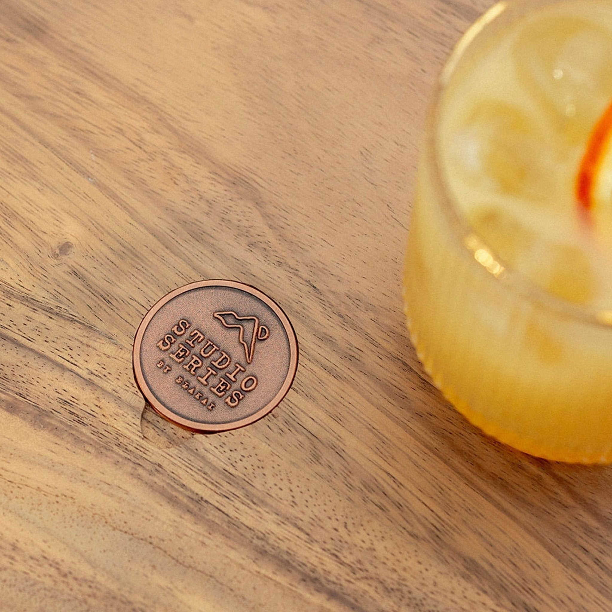 A round copper-colored metal token or coin engraved with 'AUDIO CREATION BY DAVIES' and a mountain logo, resting on a wooden tabletop next to a yellow cocktail in a glass.