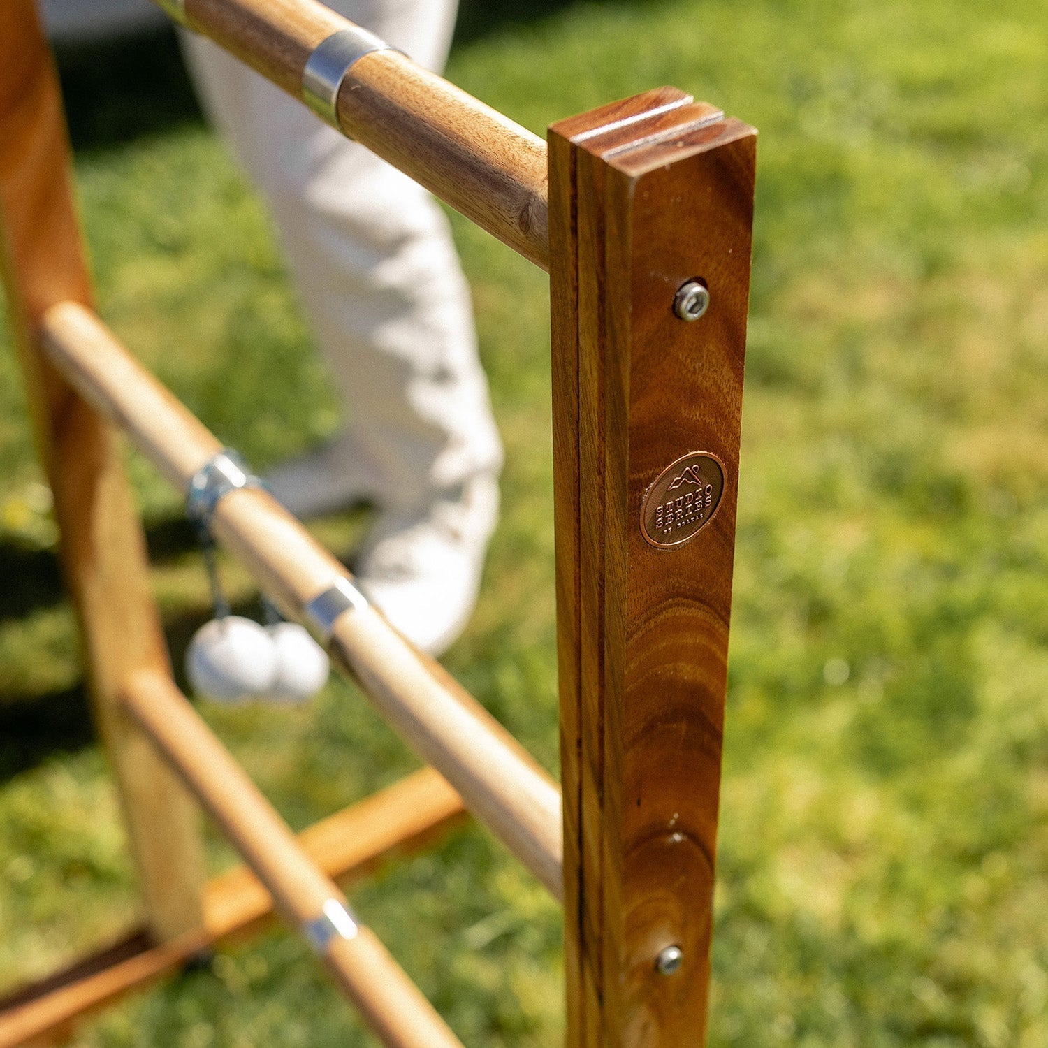 Close-up of a wooden outdoor game frame with metal connectors, shown on a grassy lawn with part of a person's legs visible in the background.