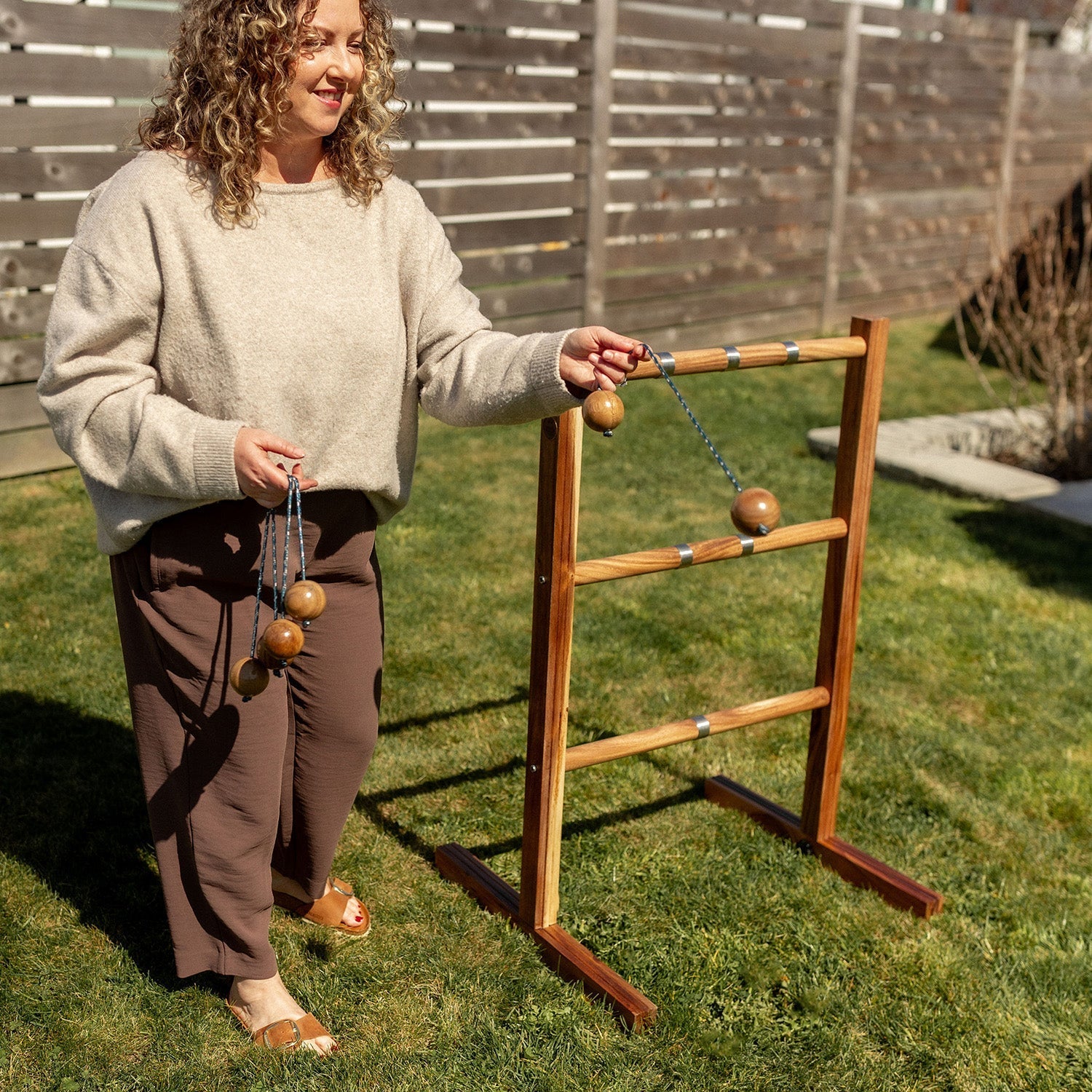 A woman stands on a grassy backyard lawn next to a wooden agility game stand with hanging wooden balls, wearing a beige sweater and brown pants.