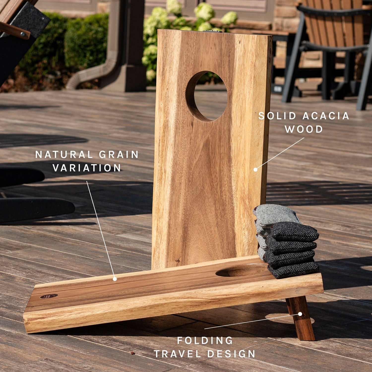 A folding wooden game board set on a deck, with text callouts reading 'Solid Acacia Wood,' 'Natural Grain Variation,' and 'Folding Travel Design.' The set includes a tall vertical post with a circular cutout and a low angled base with a circular hole, plus folded dark towels resting on the base.
