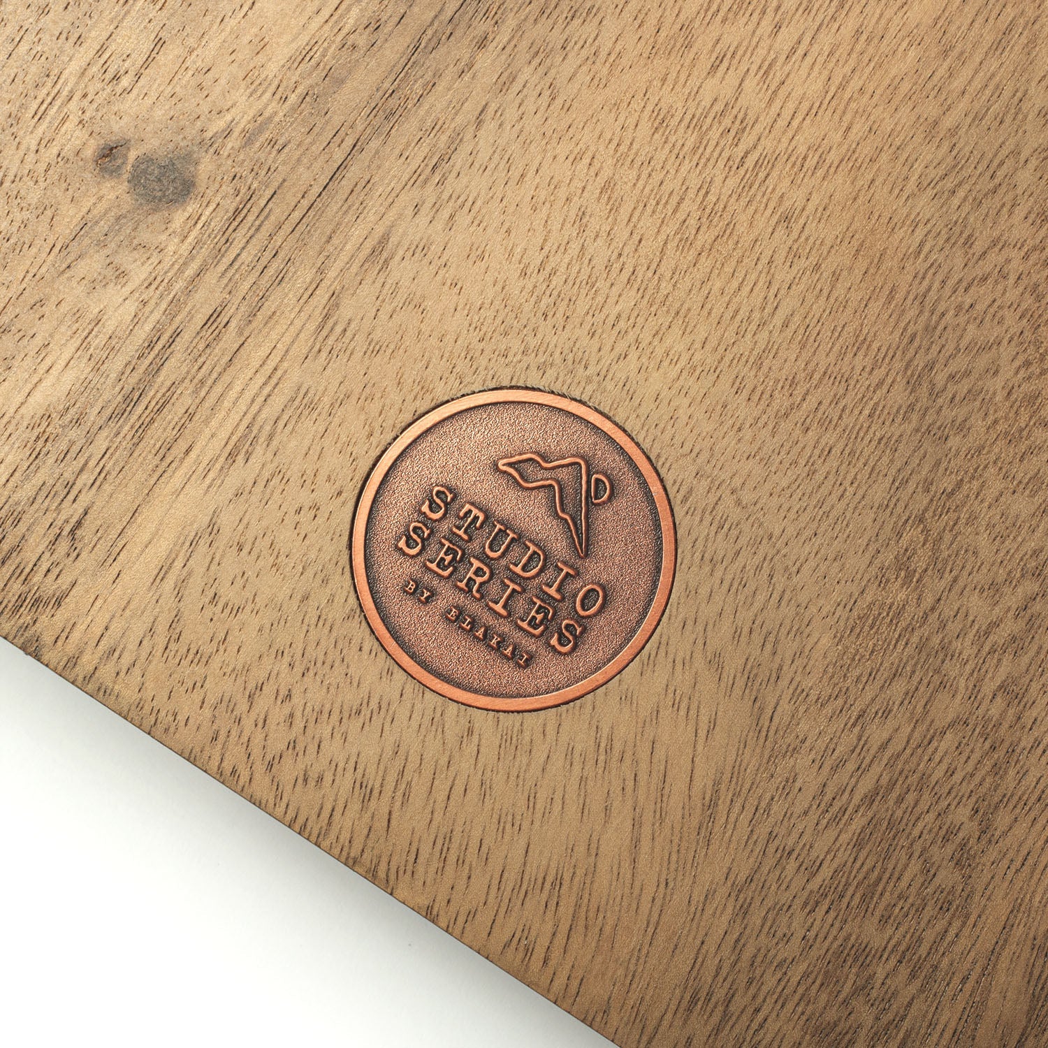 A close-up of a circular copper-toned metal emblem stamped onto a light brown wooden surface, featuring the embossed text 'STUDIO SERIES by DILLY' and a mountain-like logo.