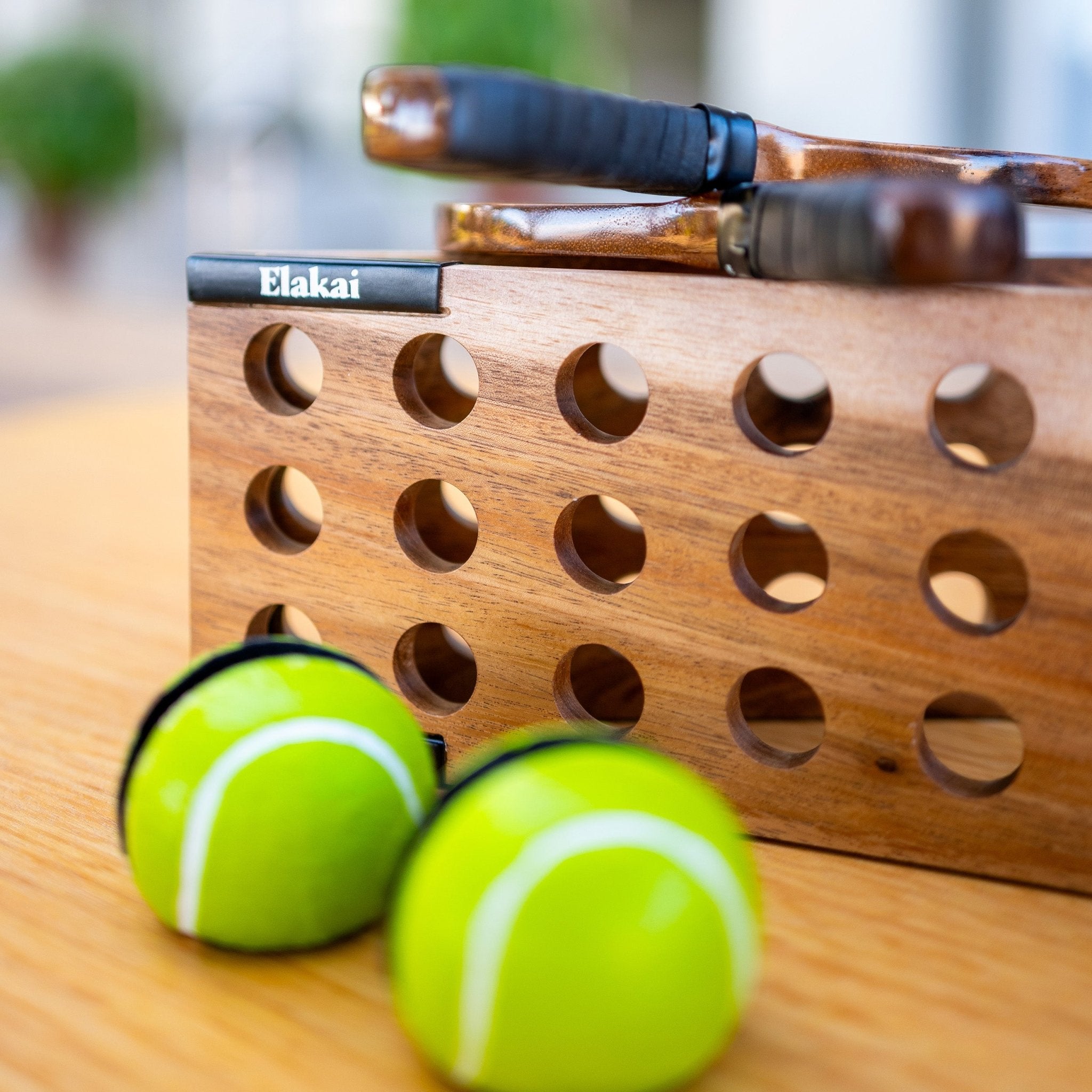 Two tennis balls and a wooden paddle holder with holes on a wooden surface.