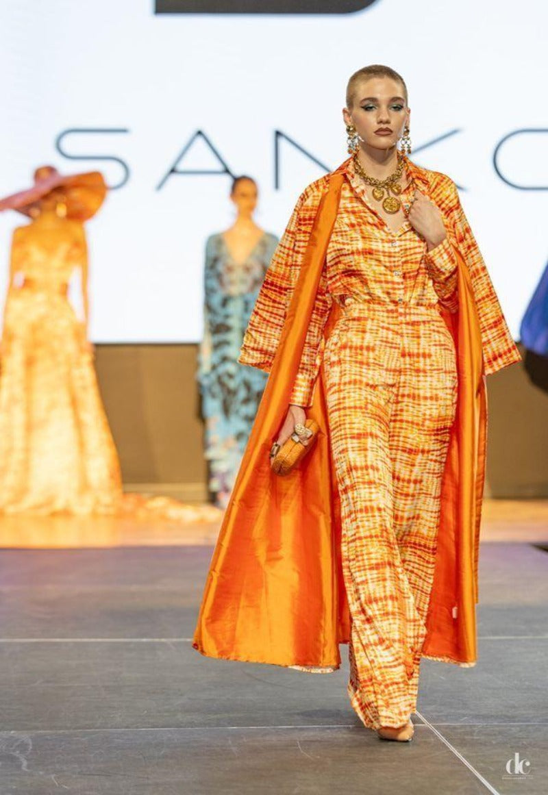Model wearing an orange and yellow outfit with a large cape at a fashion show.