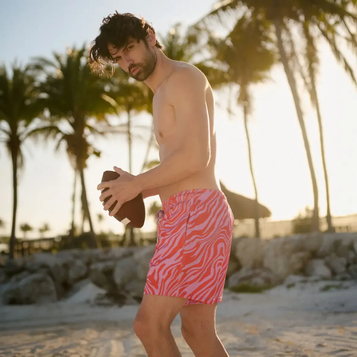 Man in pink and white zebra print swim shorts on a beach with palm trees in the background