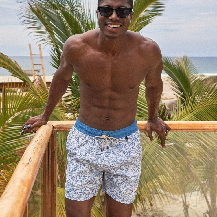 Man in swim shorts and sunglasses standing on a wooden deck with palm trees and beach in the background