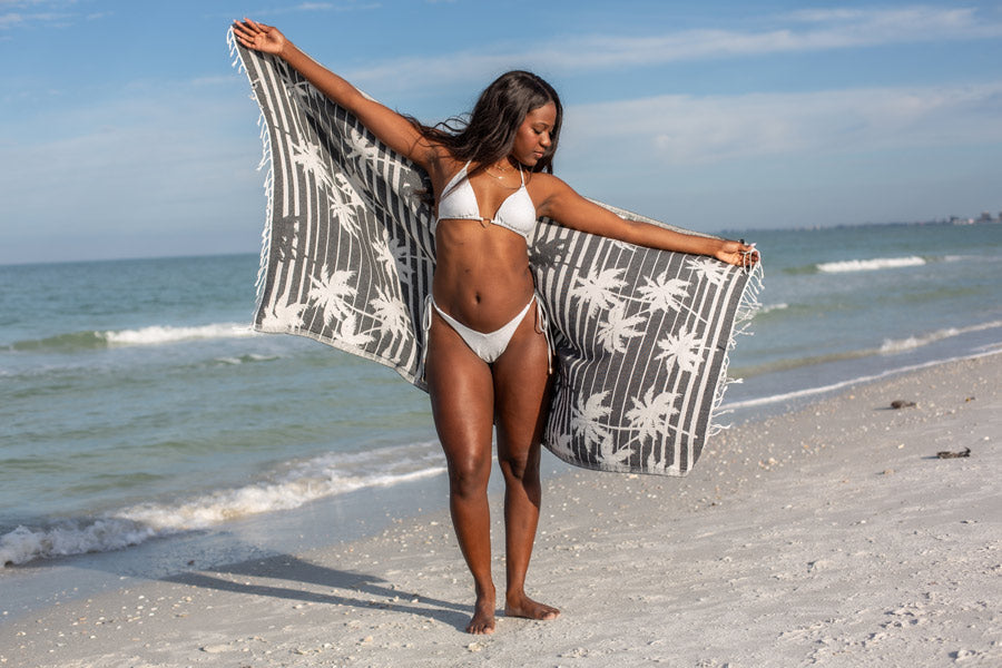 Woman on a beach holding a black and white patterned towel with palm trees.