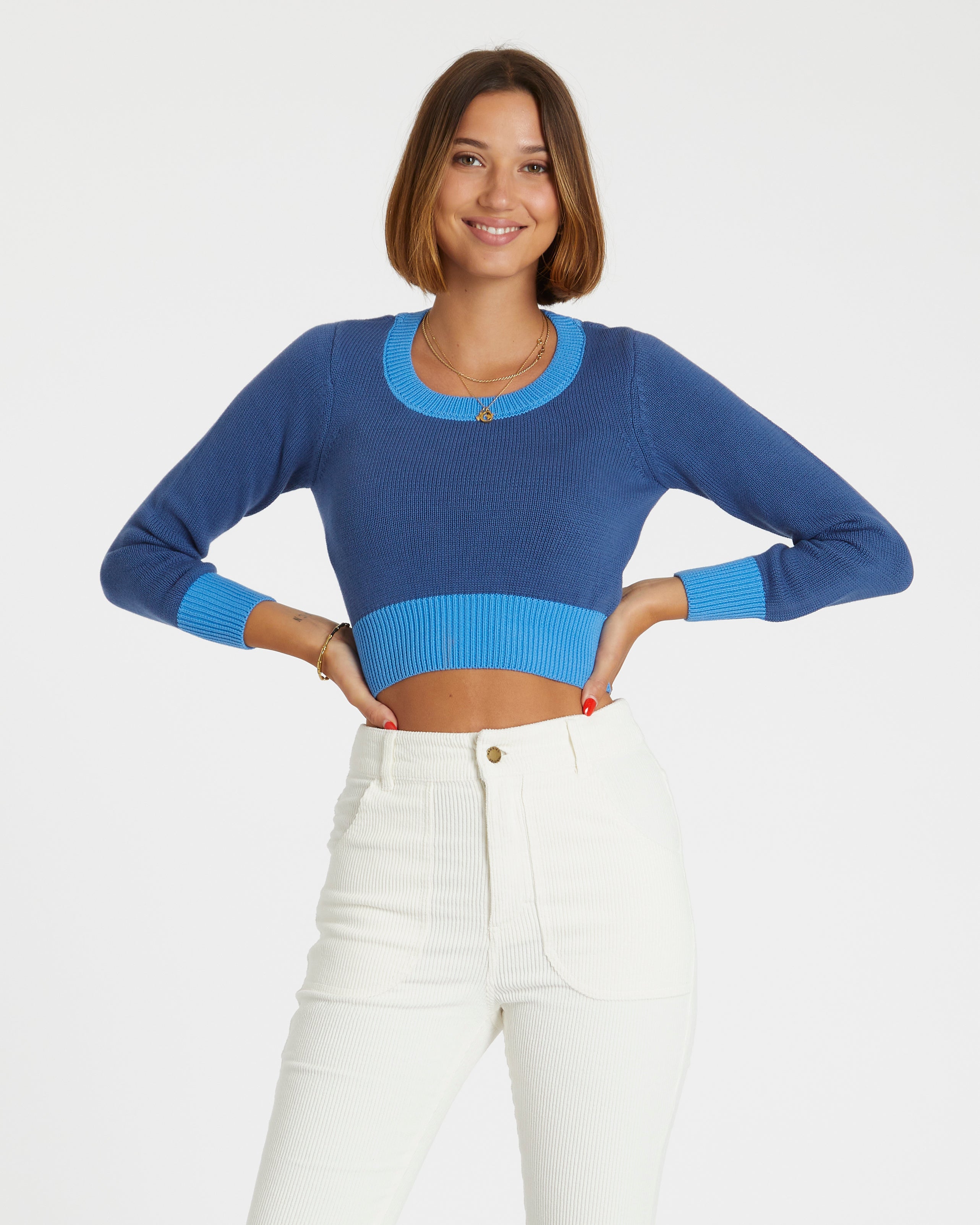 Woman wearing a blue cropped sweater and white pants on a plain background